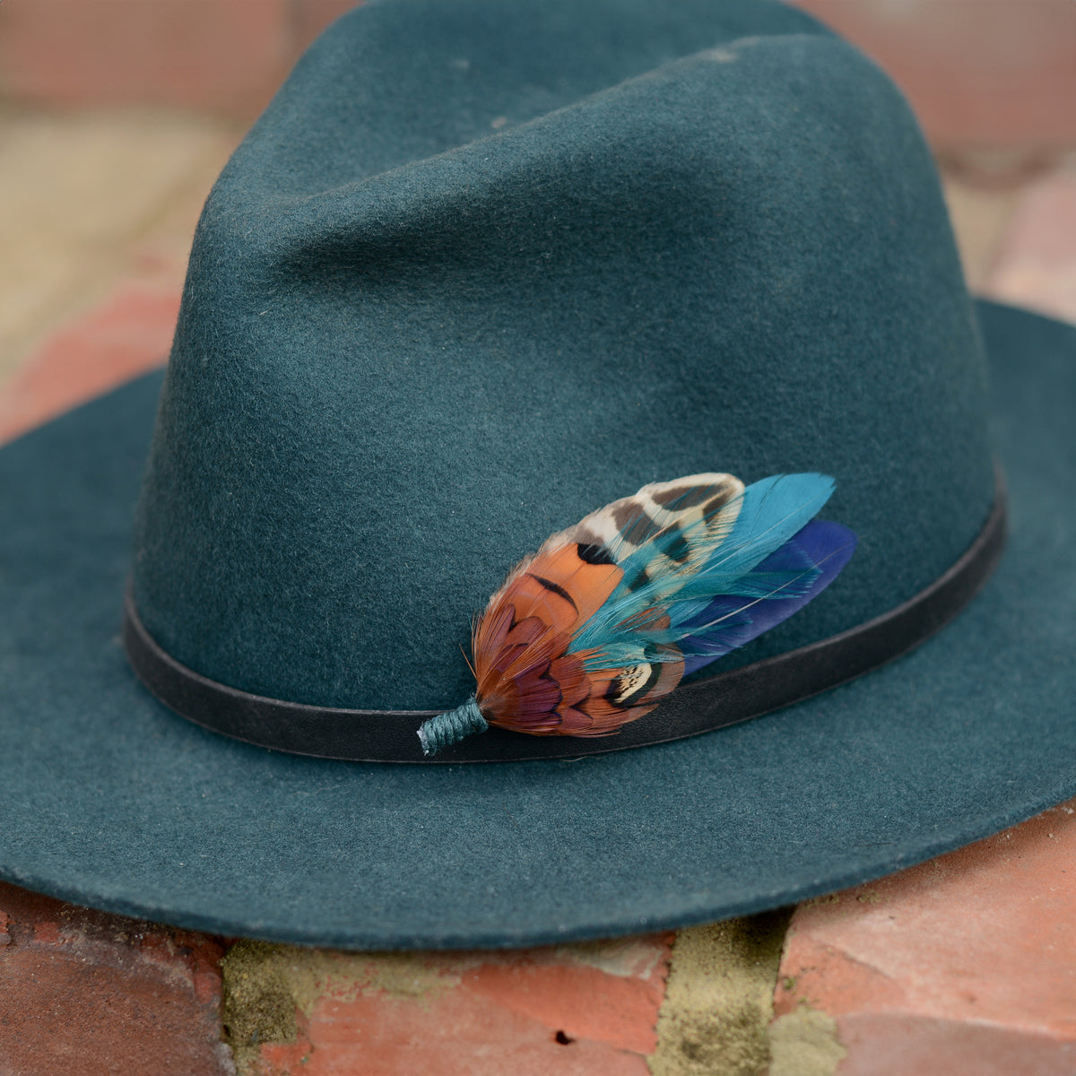 A dark teal felt fedora hat with a black band and a decorative feather hat pin in copper pheasant feathers with teal and navy, resting on a brick surface.