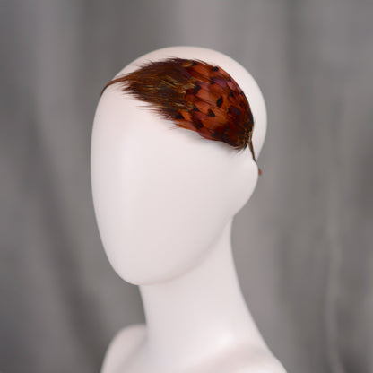 A decorative headband with a tear drop shaped pad of colourful pheasant feathers in shades of brown, orange, and purple. The feathers are attached to a thin, brown headband, creating a unique hair accessory.