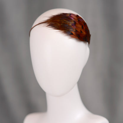A feather hair accessory featuring real natural pheasant feathers in glowing amber and copper. crafted in a layered style to hug the side of the head in a tear drop shape. Mounted on a headband covered in brown ribbon. An ideal accessory for autumn / fall weddings and countryside events