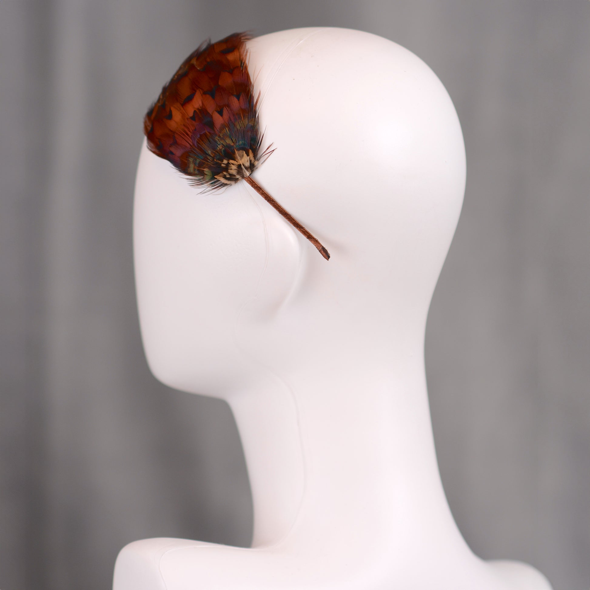 A feather hair accessory featuring real natural pheasant feathers in glowing amber and copper. crafted in a layered style to hug the side of the head in a tear drop shape. Mounted on a headband covered in brown ribbon. An ideal accessory for autumn / fall weddings and countryside events