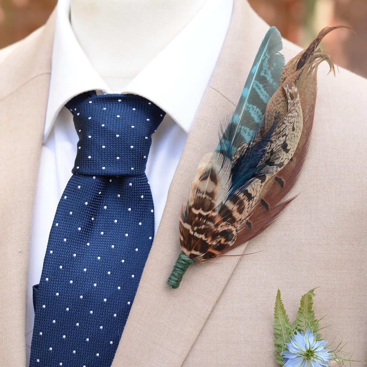 A close-up of a beige suit jacket on a mannequin torso, styled with a white dress shirt, a navy blue polka-dot tie, and a decorative feather boutonniere with brown, teal, and gold tones.