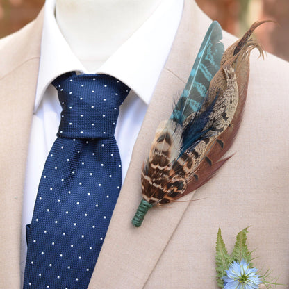 A close-up of a beige suit jacket on a mannequin torso, styled with a white dress shirt, a navy blue polka-dot tie, and a decorative feather boutonniere with brown, teal, and gold tones.