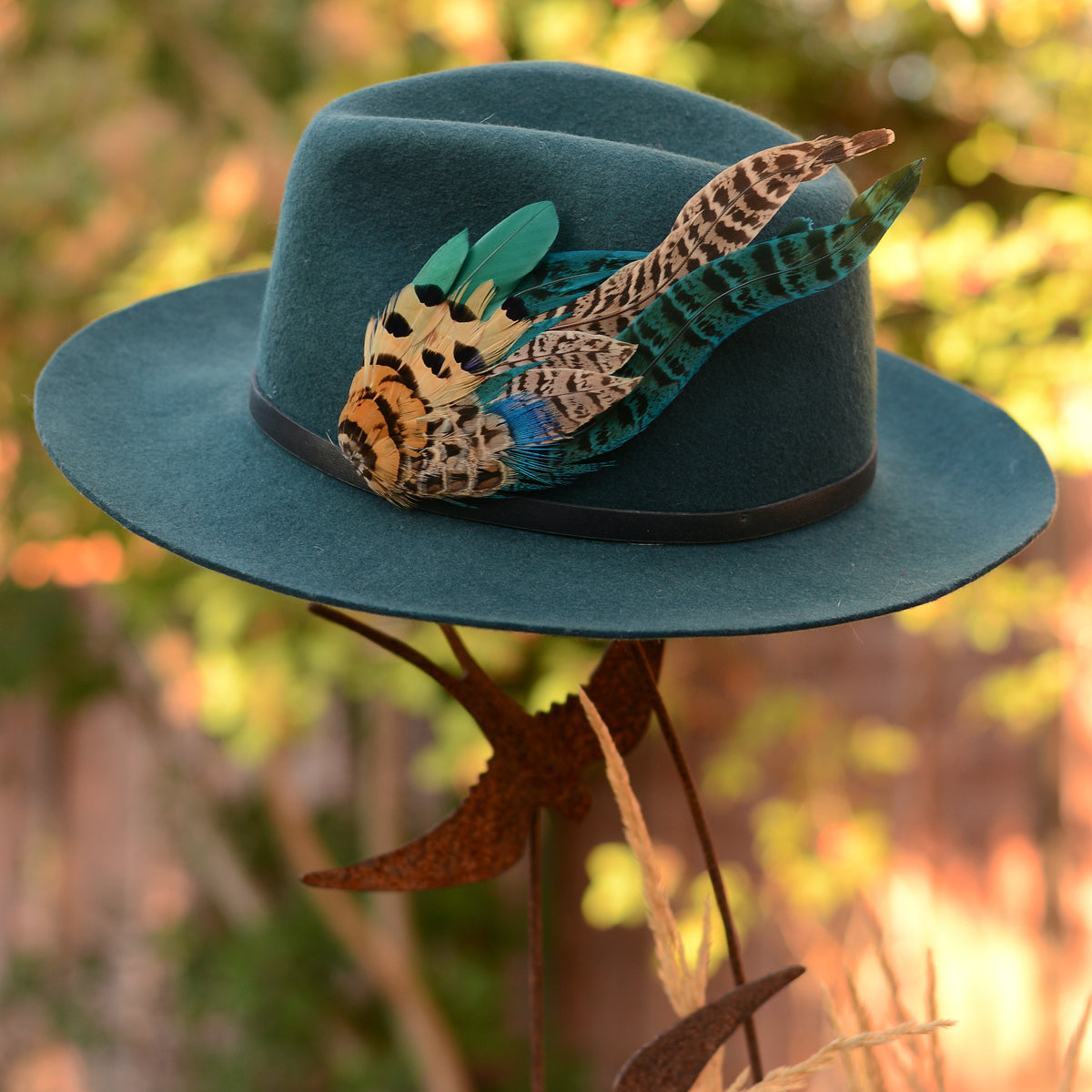 A teal blue felt wide-brim hat decorated with a large multicolour feather hat pin embellishment, displayed on a metal stand against a blurred outdoor background.
