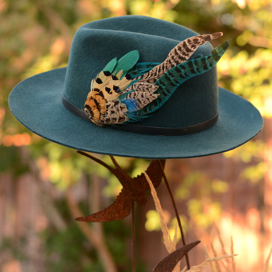 A teal blue felt wide-brim hat decorated with a large multicolour feather hat pin embellishment, displayed on a metal stand against a blurred outdoor background.
