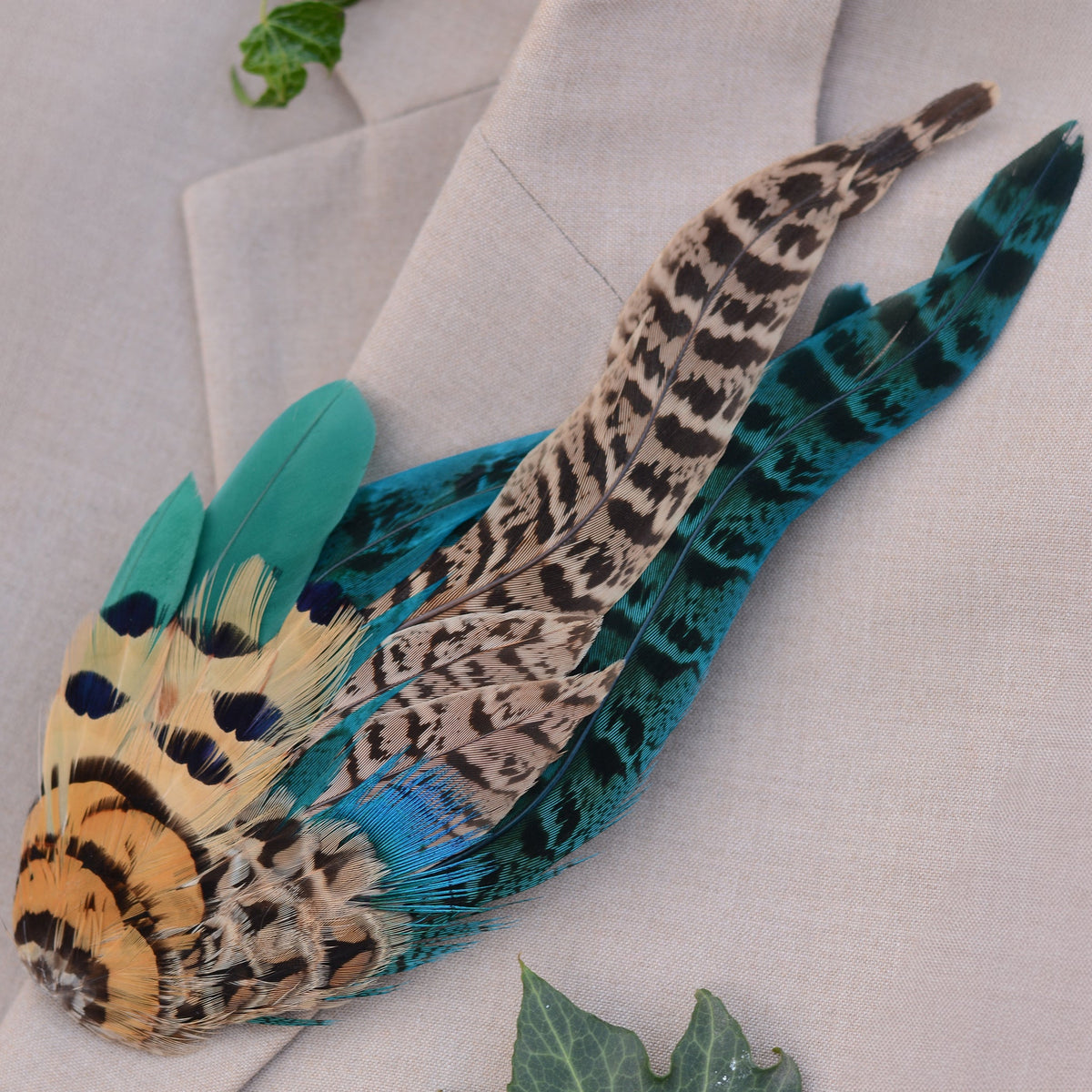 A decorative feather pin or hat accessory with layered pheasant-style feathers in teal, black, tan, and gold, arranged on a light beige fabric background with small green leaves.