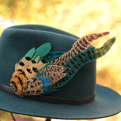 A dark teal felt cowboy hat with a black band and a large decorative hat pin in the style of a feather plume in teal, tan, black, and brown, photographed against a warm golden bokeh background.