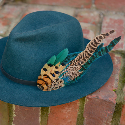 A teal blue felt wide-brim hat with a black band and decorative feather hat pin in teal, tan, black, and blue, resting on a brick surface.