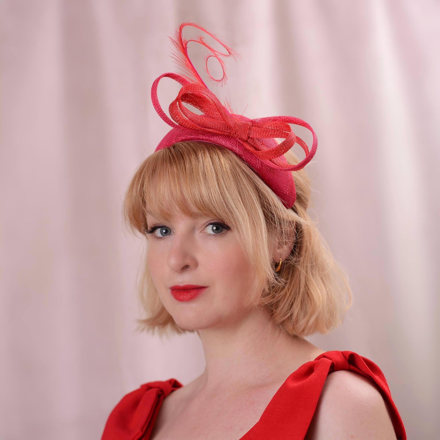 A woman with short blonde hair wearing a bright pink and red bandeau fascinator hat with a bow and curled feathers paired and a red dress, photographed against a soft pink curtain background.