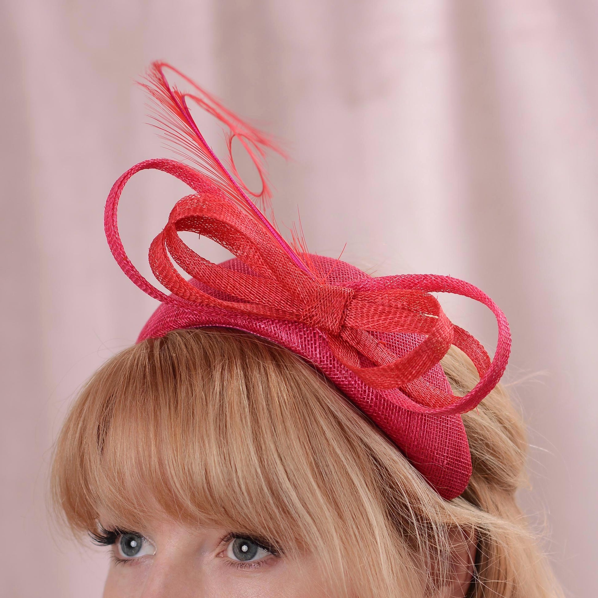 A close-up of a woman wearing a bright pink and red fascinator headband with a large bow and feather, set against a soft pink background.