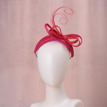A bright pink and red Royal Ascot fascinator hat with a large bow and curling feather detail, displayed on a white mannequin head against a soft pink background.