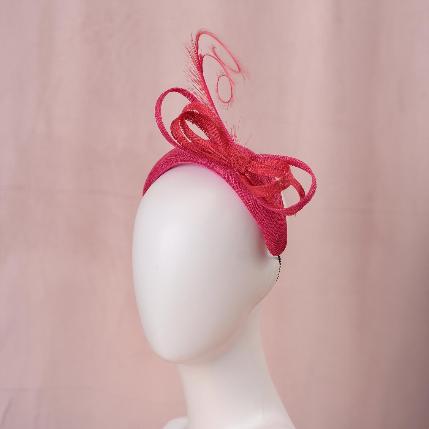 A bright pink and red horse racing headpiece hat with a large bow and curling feather detail, displayed on a white mannequin head against a soft pink background.