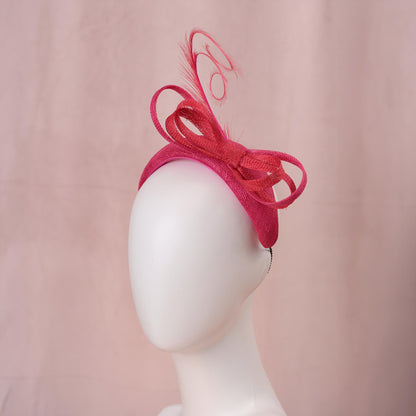 A bright pink and red horse racing headpiece hat with a large bow and curling feather detail, displayed on a white mannequin head against a soft pink background.