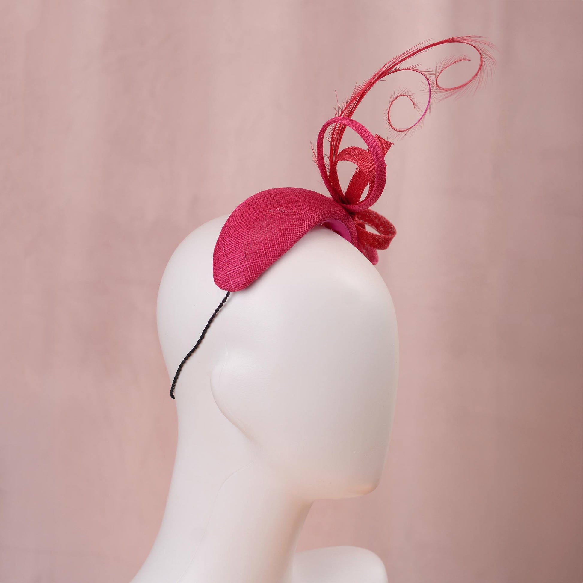 A bright pink and red horse racing headband hat with a large bow and curling feather detail, displayed on a white mannequin head against a soft pink background.