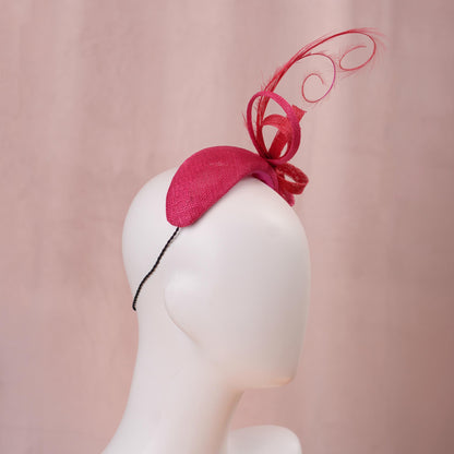 A bright pink and red horse racing headband hat with a large bow and curling feather detail, displayed on a white mannequin head against a soft pink background.