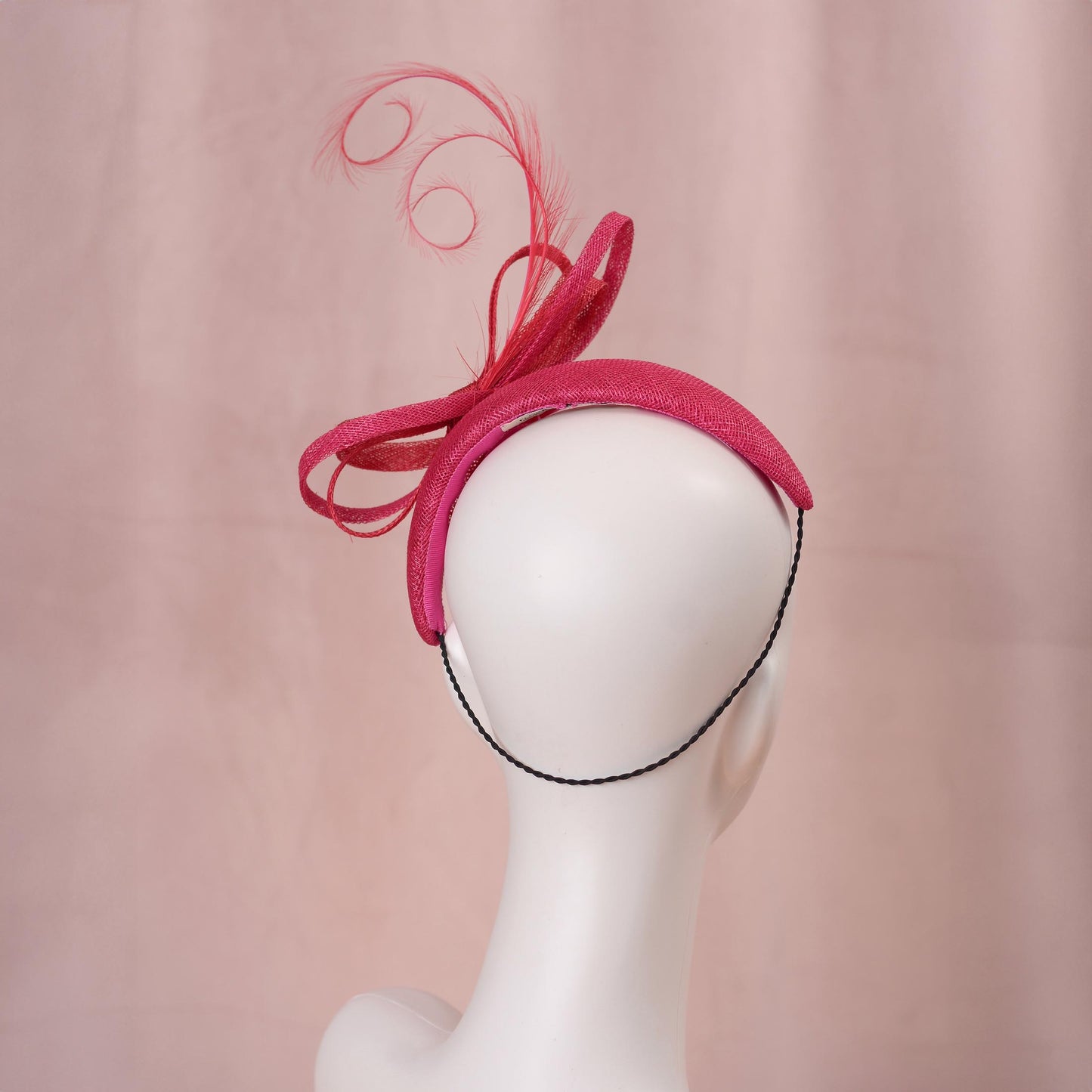 A bright pink and red horse racing headband hat with a large bow and curling feather detail, displayed on a white mannequin head against a soft pink background.