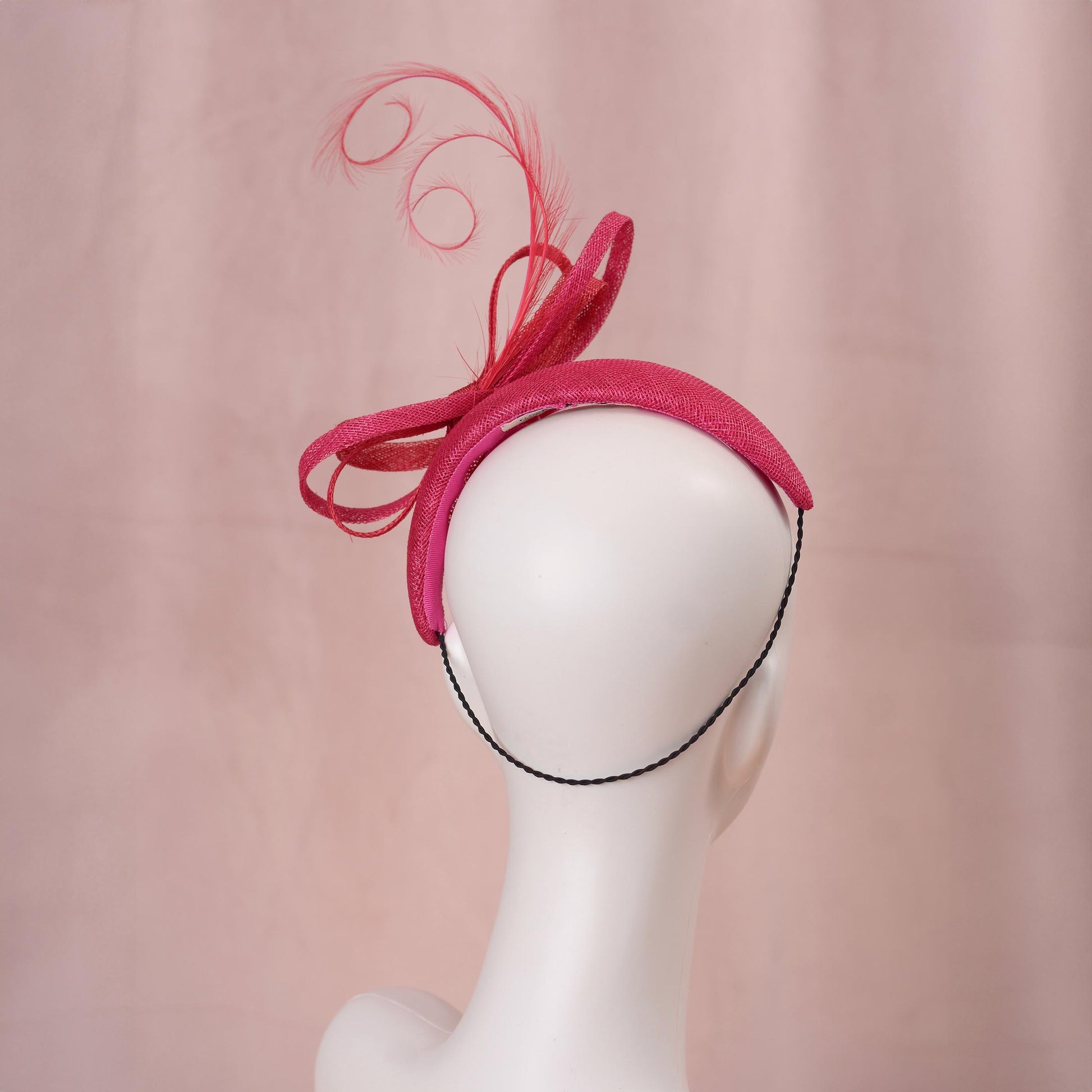 A bright pink and red horse racing headband hat with a large bow and curling feather detail, displayed on a white mannequin head against a soft pink background.