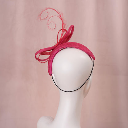 A bright pink and red horse racing headband hat with a large bow and curling feather detail, displayed on a white mannequin head against a soft pink background.