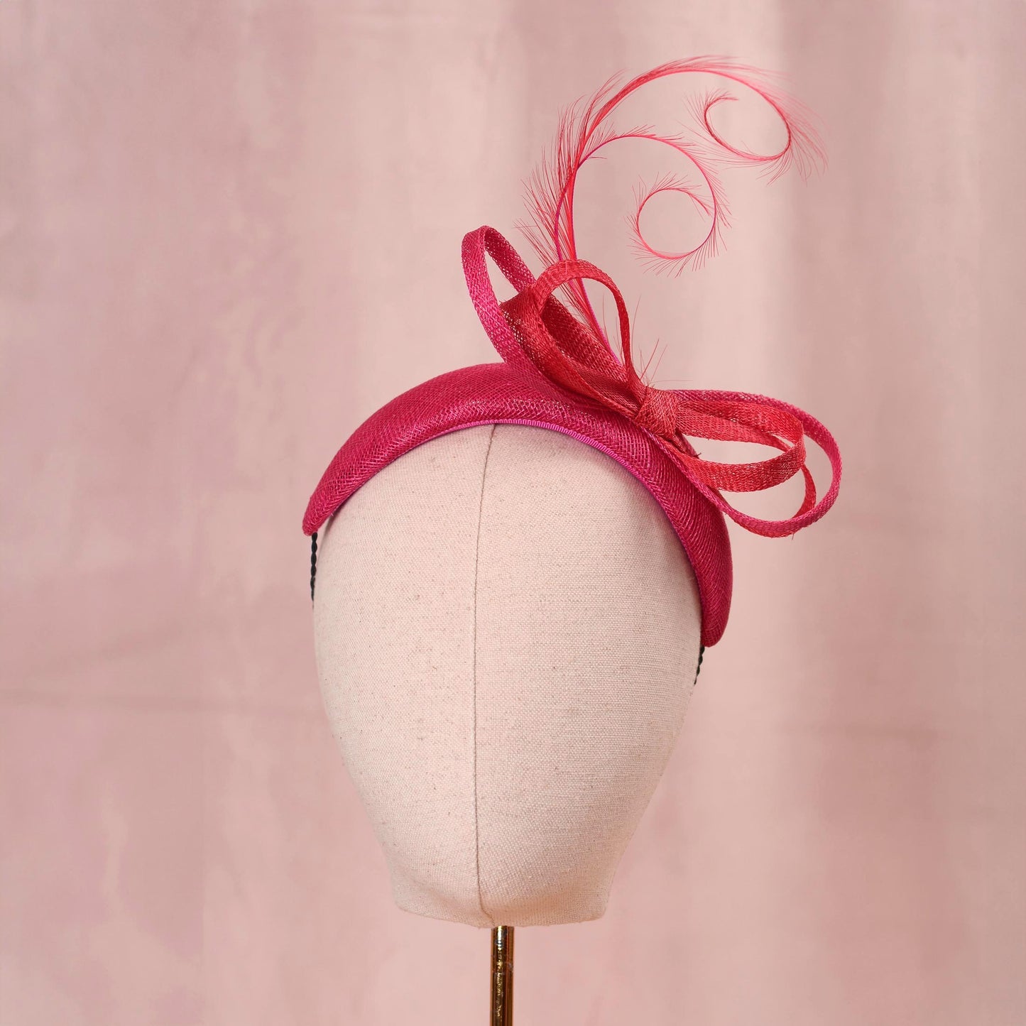 A bright pink and wedding headpiece hat with a large bow and curling feather detail, displayed on a white mannequin head against a soft pink background.
