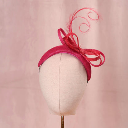 A bright pink and wedding headpiece hat with a large bow and curling feather detail, displayed on a white mannequin head against a soft pink background.