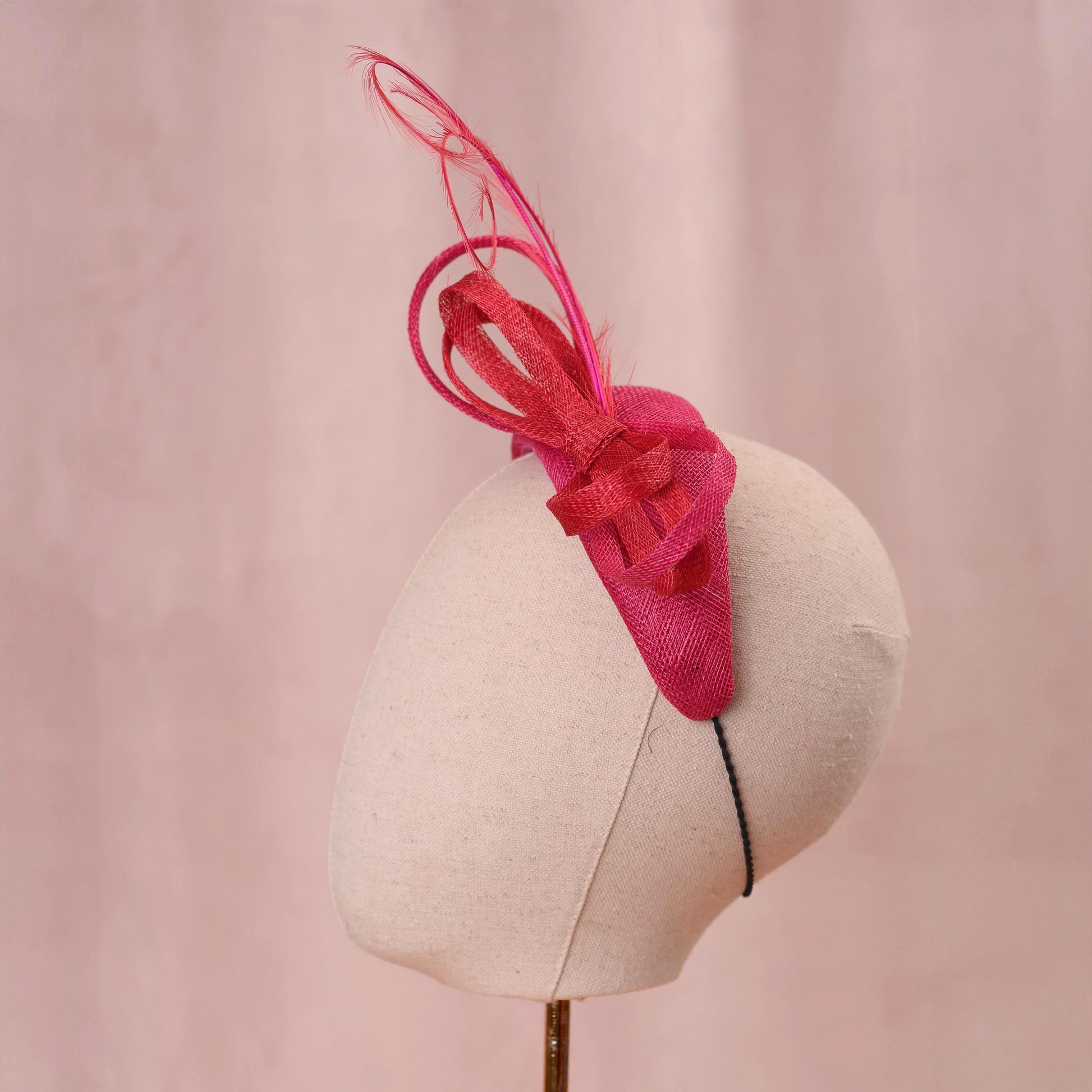 A bright pink and red horse racing headband hat with a large bow and curling feather detail, displayed on a white mannequin head against a soft pink background.