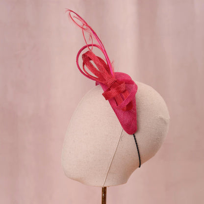A bright pink and red horse racing headband hat with a large bow and curling feather detail, displayed on a white mannequin head against a soft pink background.