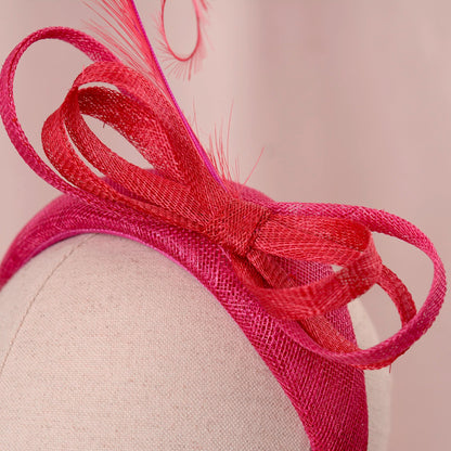close up of a bright pink and red horse racing headband hat with a large bow and curling feather detail, displayed on a white mannequin head against a soft pink background.