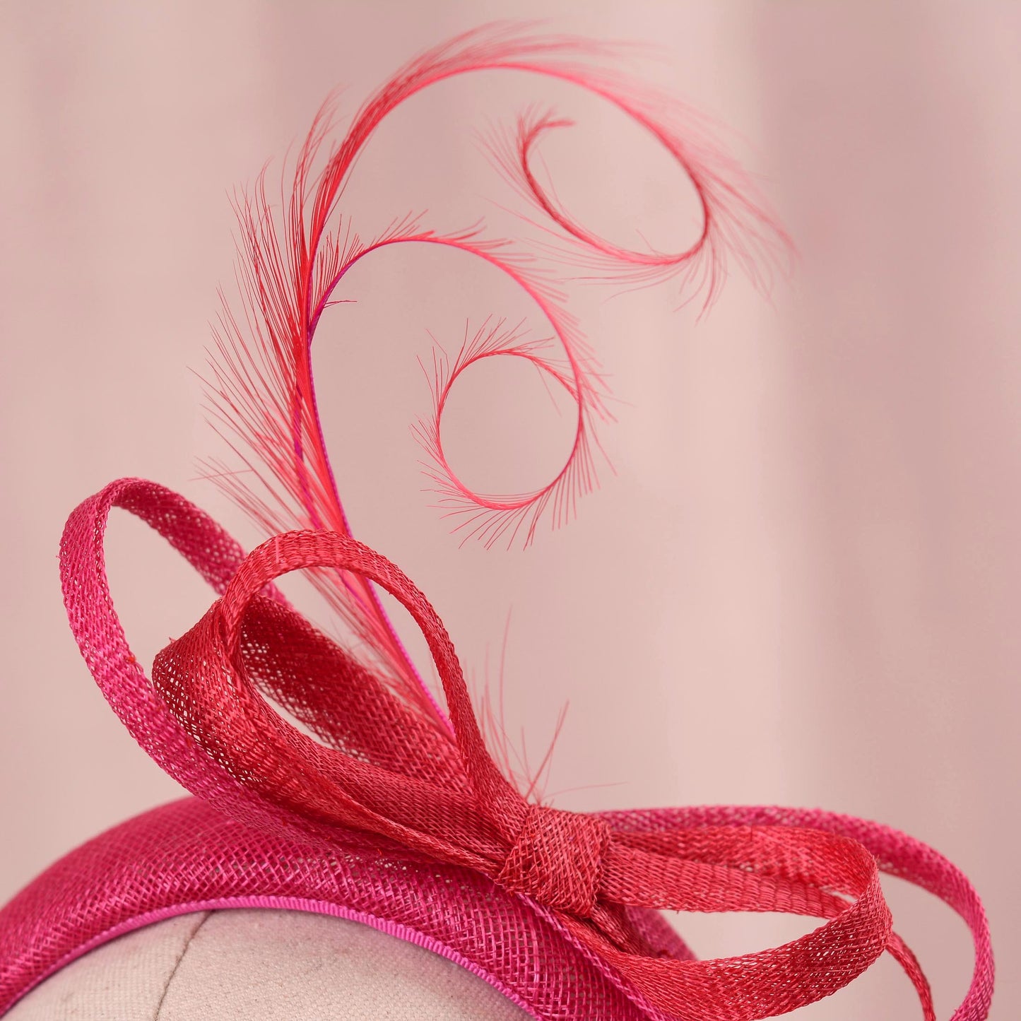 A close-up of a bright pink fascinator hat with a large sinamay bow and a tall feather plume on a mannequin head against a soft pink background.