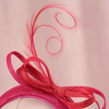 A close-up of a bright pink fascinator hat with a large sinamay bow and a tall feather plume on a mannequin head against a soft pink background.