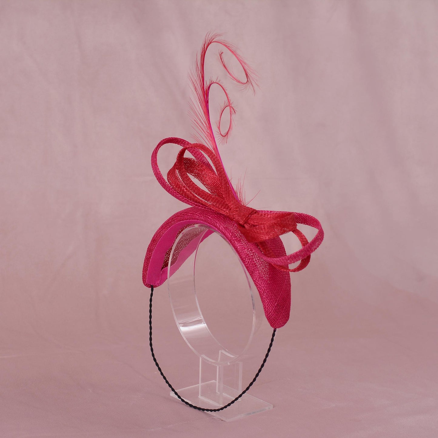 A bright pink and red fascinator hat with a large bow and feather accent, displayed on a clear plastic stand against a pink fabric backdrop.