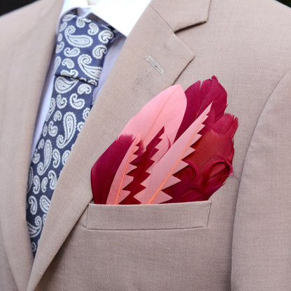 A close-up of a light beige linen jacket pocket holding a decorative pocket square featuring a fan of goose feathers in shades of pink and deep magenta-red cut into graphic shapes to produce contrast and curled for texture.