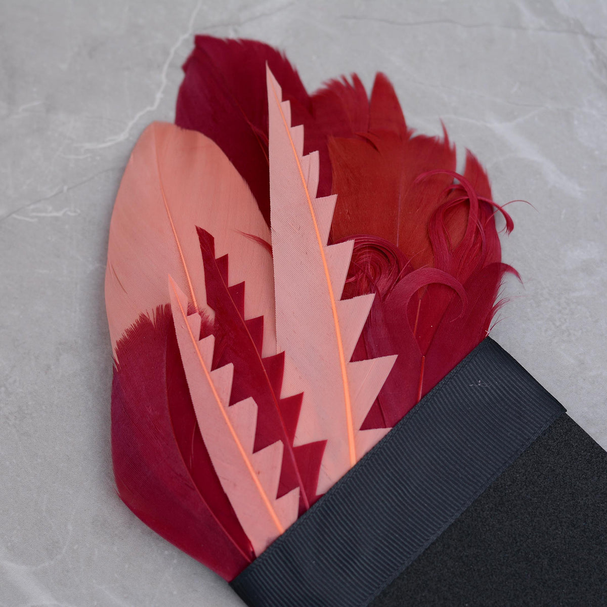 A decorative pocket square featuring a fan of goose feathers in shades of pink and deep magenta-red cut into graphic shapes to produce contrast and curled for texture. Laid on a grey stone background.