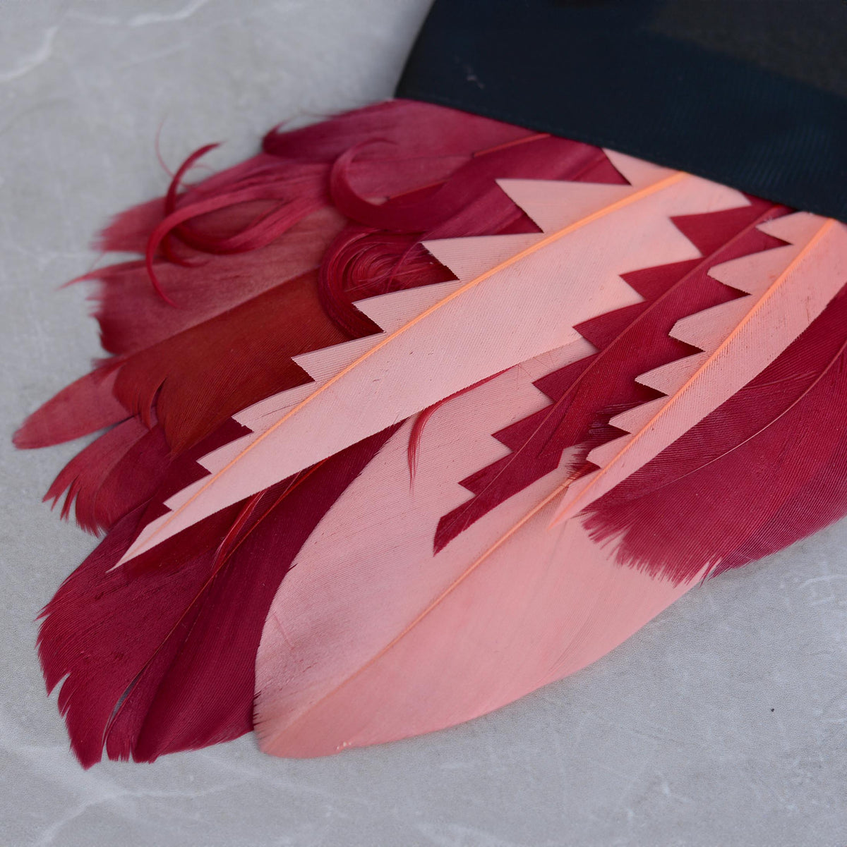 A decorative pocket square featuring a fan of goose feathers in shades of pink and deep magenta-red cut into graphic shapes to produce contrast and curled for texture. Laid on a grey stone background.