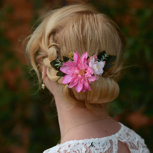 A blonde woman shown from the back wearing a floral hair accessory with a large pink flower, a smaller pale pink /white flower, and green leaves with  her hair in a updo.