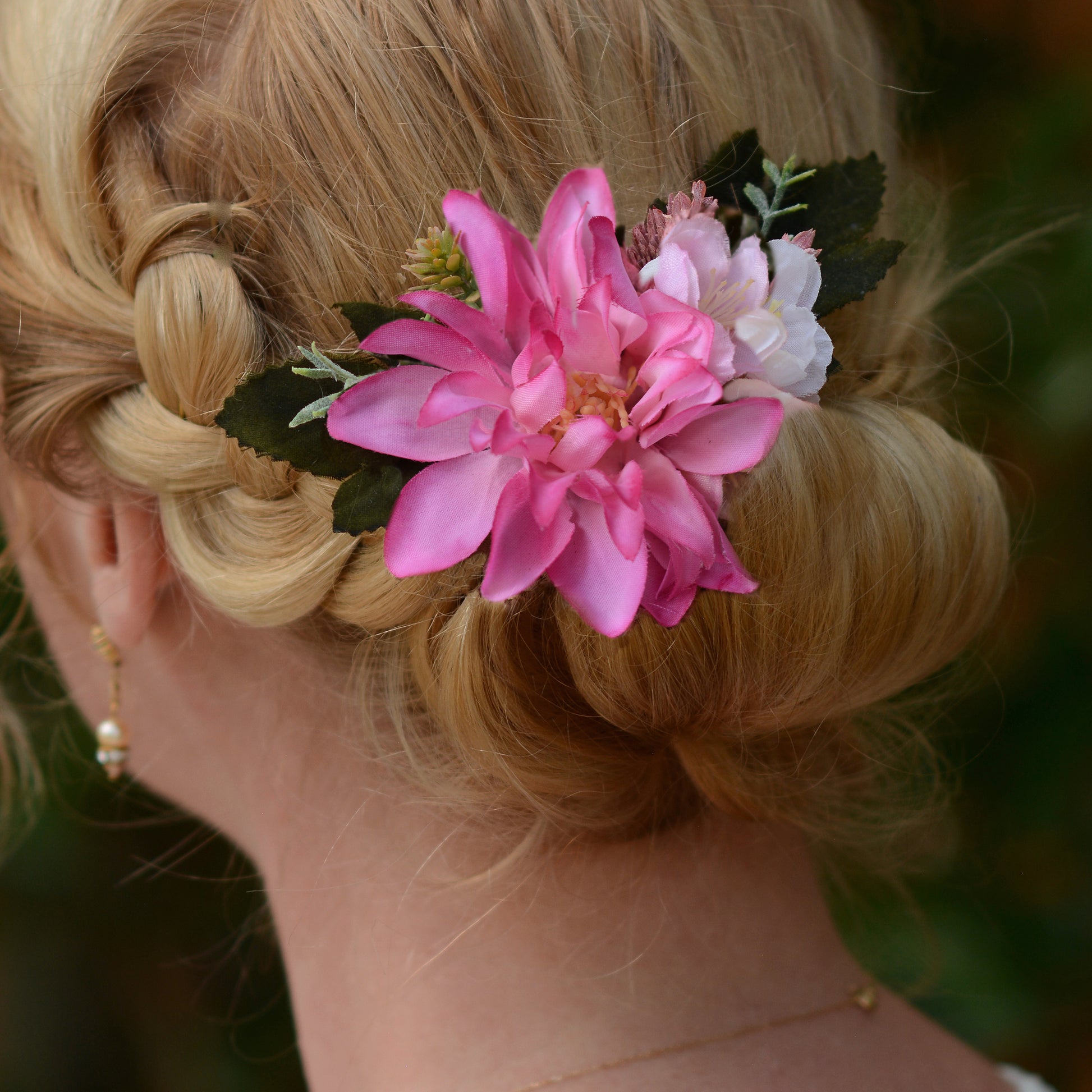 Close up of a blonde woman shown from the back wearing a floral hair accessory with a large pink flower, a smaller pale pink /white flower, and green leaves with  her hair in a updo.