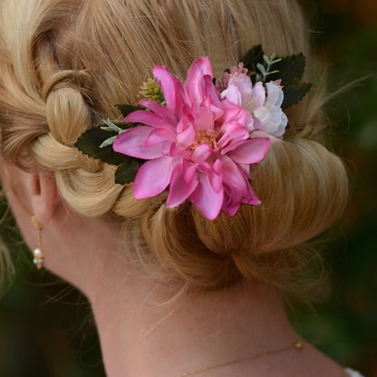 Close up of a blonde woman shown from the back wearing a floral hair accessory with a large pink flower, a smaller pale pink /white flower, and green leaves with  her hair in a updo.