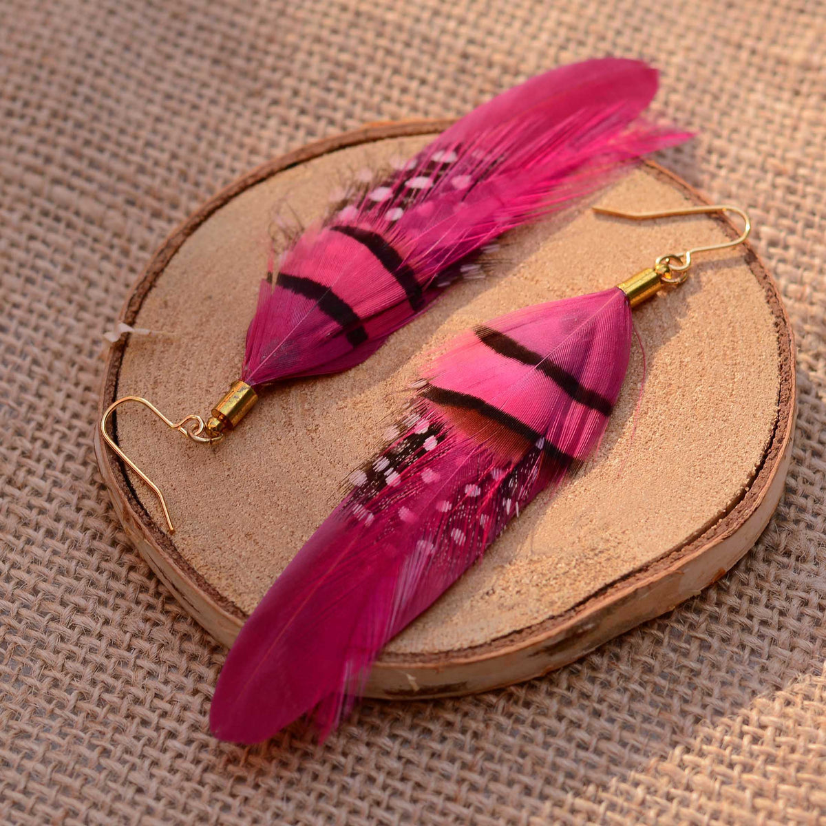 A pair of long dangling feather earrings in bright pink and magenta tones with black bands and small white speckles, attached to gold-tone fishhook wires, displayed on a round wood slice over a burlap background.