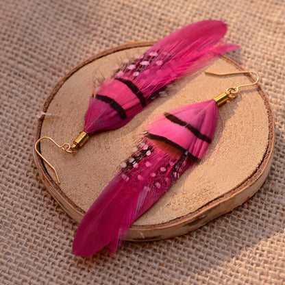 A pair of long dangling feather earrings in bright pink and magenta tones with black bands and small white speckles, attached to gold-tone fishhook wires, displayed on a round wood slice over a burlap background.