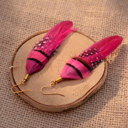 A pair of bohemian-style feather earrings with vibrant pink ombre-effect feathers, black stripes, and white speckles, hanging from gold fishhook wires on a round wooden slice over a burlap background.