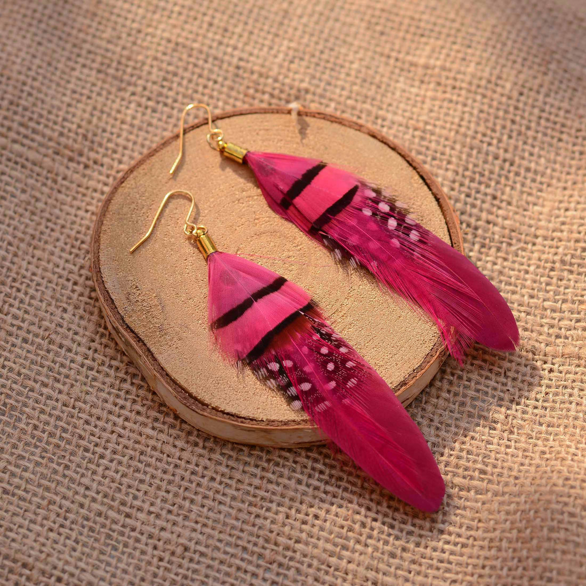 A pair of dangling feather festival earrings in bright pink and magenta pink tones with black and white speckled accents, displayed on a round wood slice over a burlap fabric background.