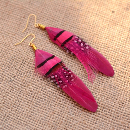 A pair of long feather drop earrings in bright pink and magenta tones with black and white speckled accents, hanging from gold fishhook earwires on a textured beige burlap background.