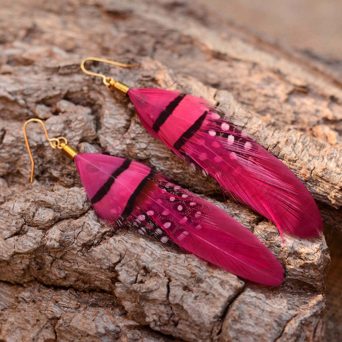 A pair of dangling bohemian feather earrings with bright pink feathers, black bands, and small white dot details, attached to gold fishhook wires, displayed on a textured brown bark surface.