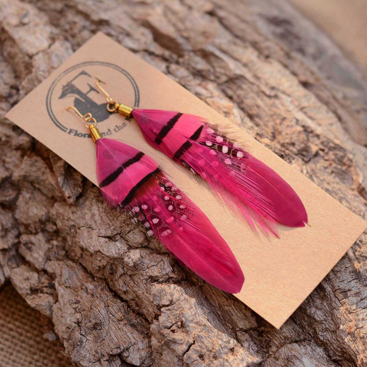 A pair of long feather dangle earrings in vivid pink with black stripes and small white dot details, displayed on a kraft paper card with a logo, resting on a textured tree bark surface.