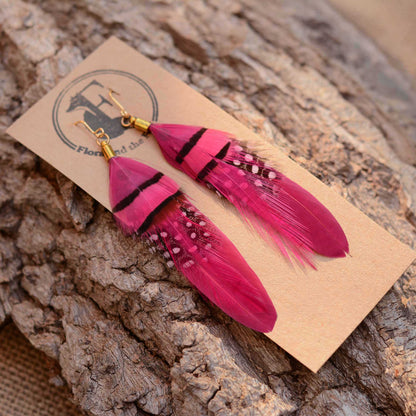 A pair of long feather dangle earrings in vivid pink with black stripes and small white dot details, displayed on a kraft paper card with a logo, resting on a textured tree bark surface.