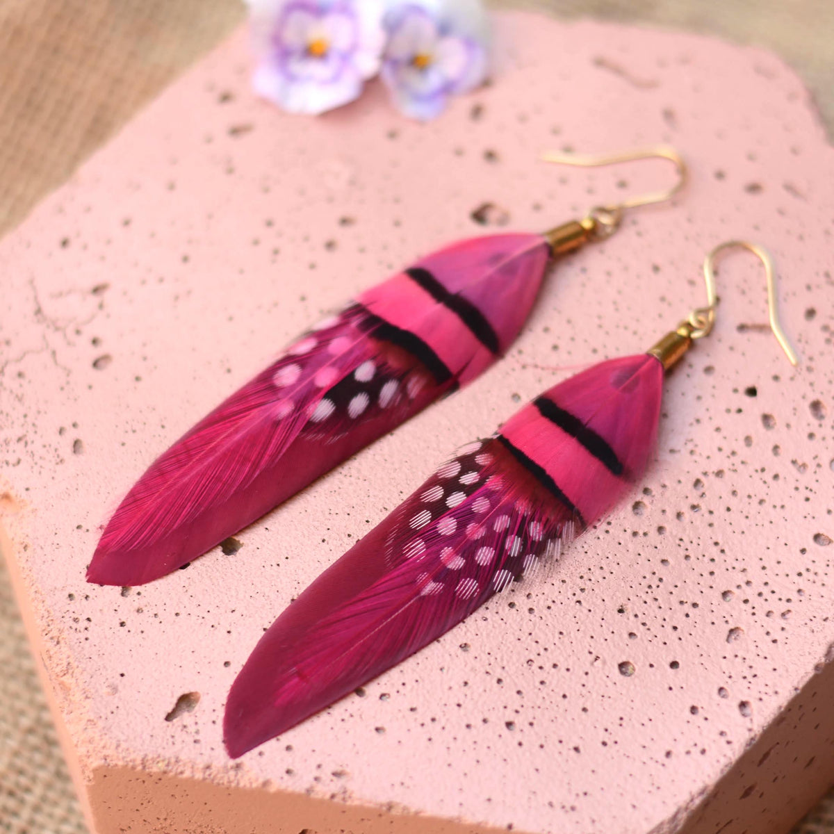A pair of long festival feather drop earrings in vibrant magenta pink and deep raspberry, featuring black stripes, white spots, and gold fishhook wires, displayed on a textured pink stone surface.