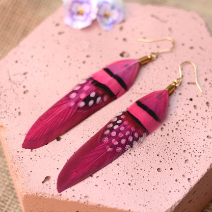 A pair of long festival feather drop earrings in vibrant magenta pink and deep raspberry, featuring black stripes, white spots, and gold fishhook wires, displayed on a textured pink stone surface.