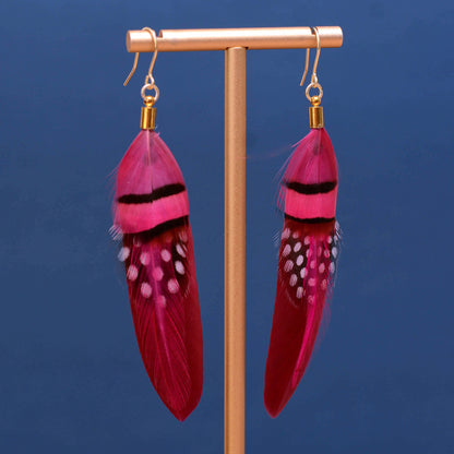 A pair of long bright pink feather drop earrings displayed on a metallic stand against a solid dark blue background. The earrings feature vibrant magenta-pink feathers with black bands and white spots, suspended from gold fishhook wires with gold caps.