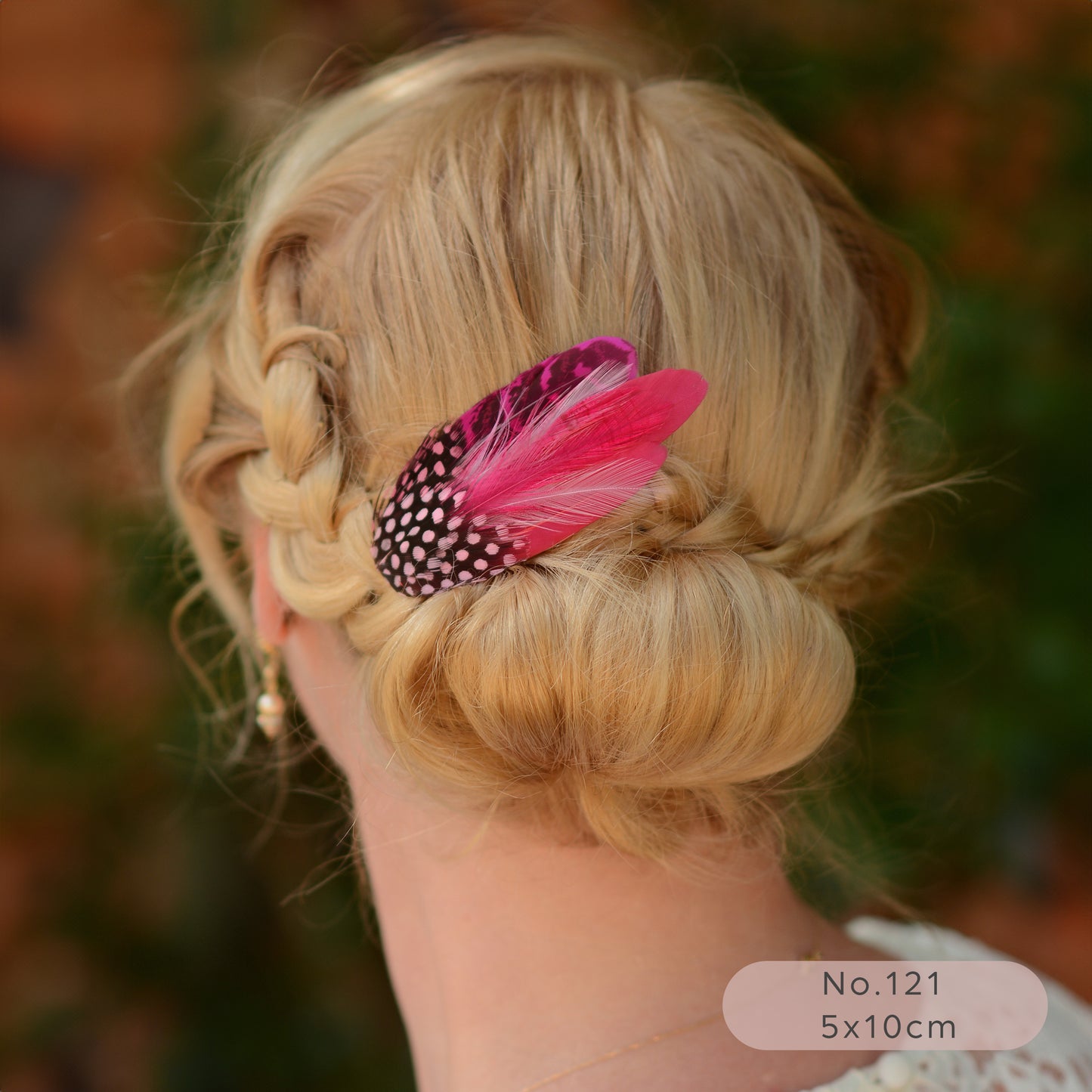 Blonde hair styled in an updo with a bright pink feather hair  accessory, blurred natural background