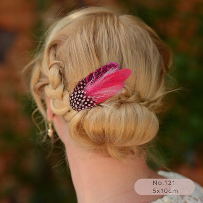 Blonde hair styled in an updo with a bright pink feather hair  accessory, blurred natural background