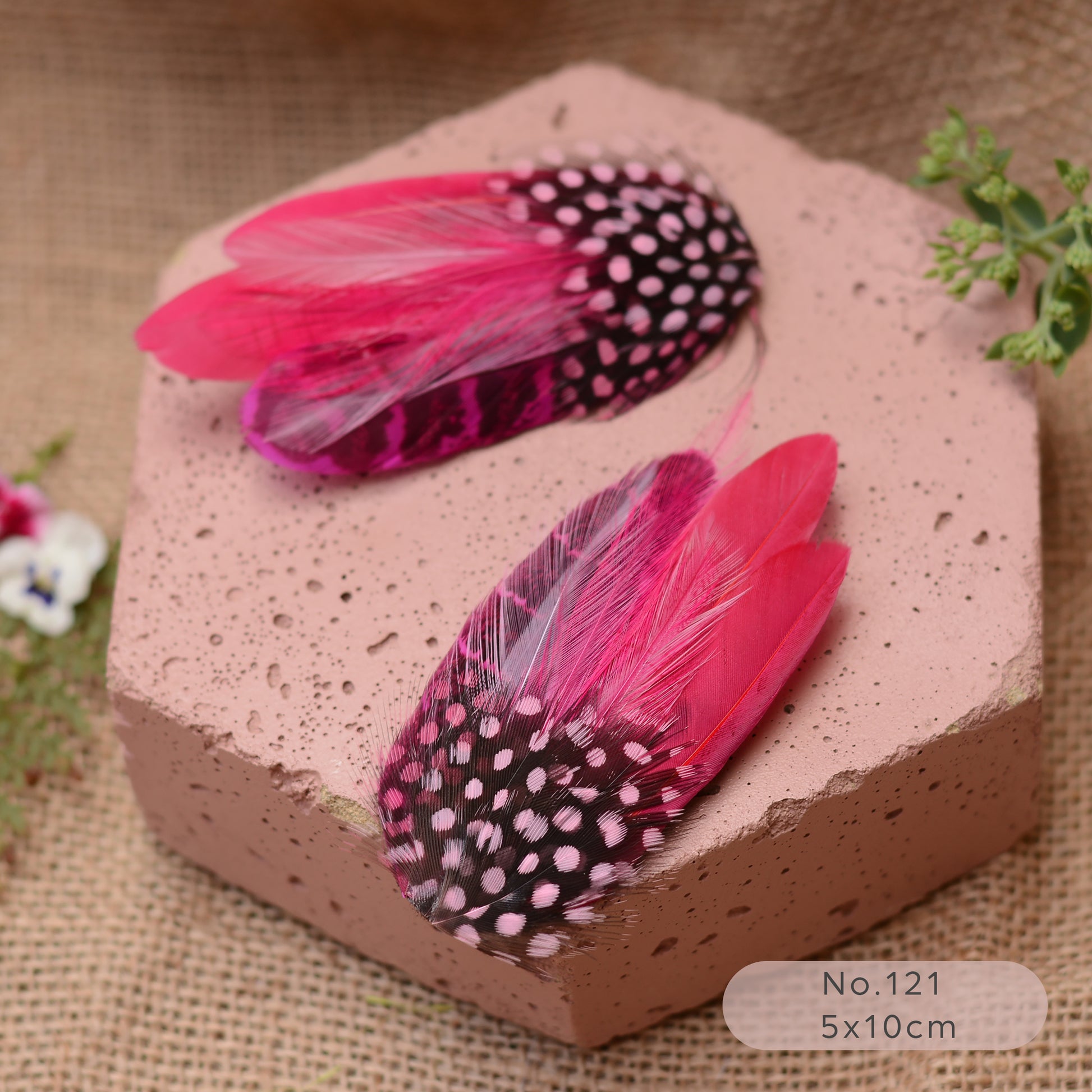 Pink and black spotted feathered hair clips on a textured pink block with a burlap background.
