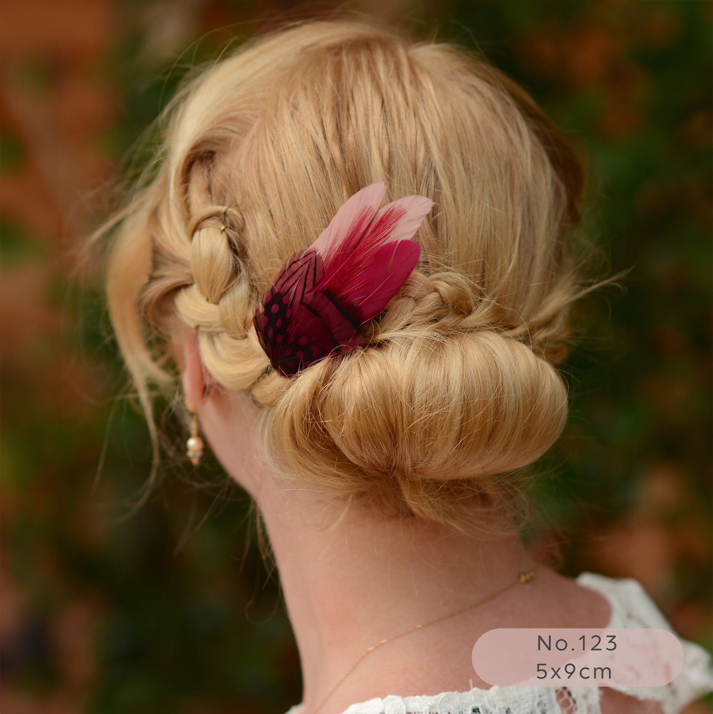 Blonde hair styled in an updo with a pink and burgundy feather accessory, blurred natural background